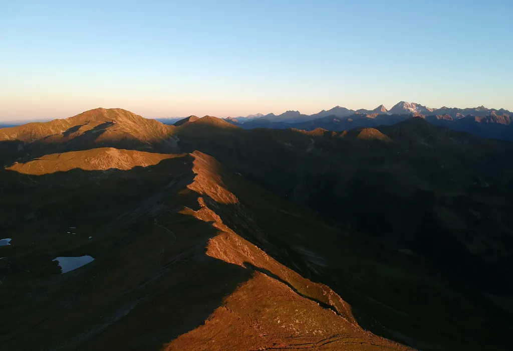 A grassy alpine ridge lit orange by low sun, with distant peaks under a clear sky