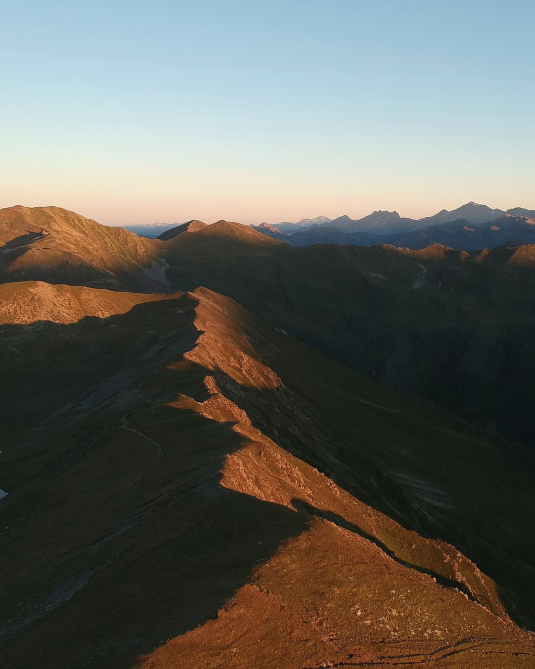 A grassy alpine ridge lit orange by low sun, with distant peaks under a clear sky