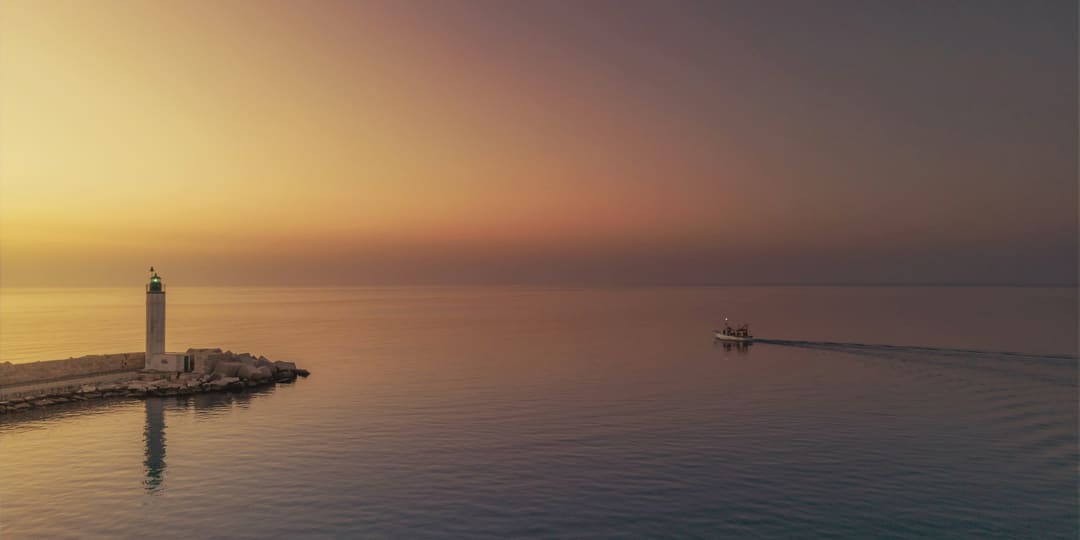 The low sun over a calm sea, with a lighthouse at the end of a harbour wall and one small boat