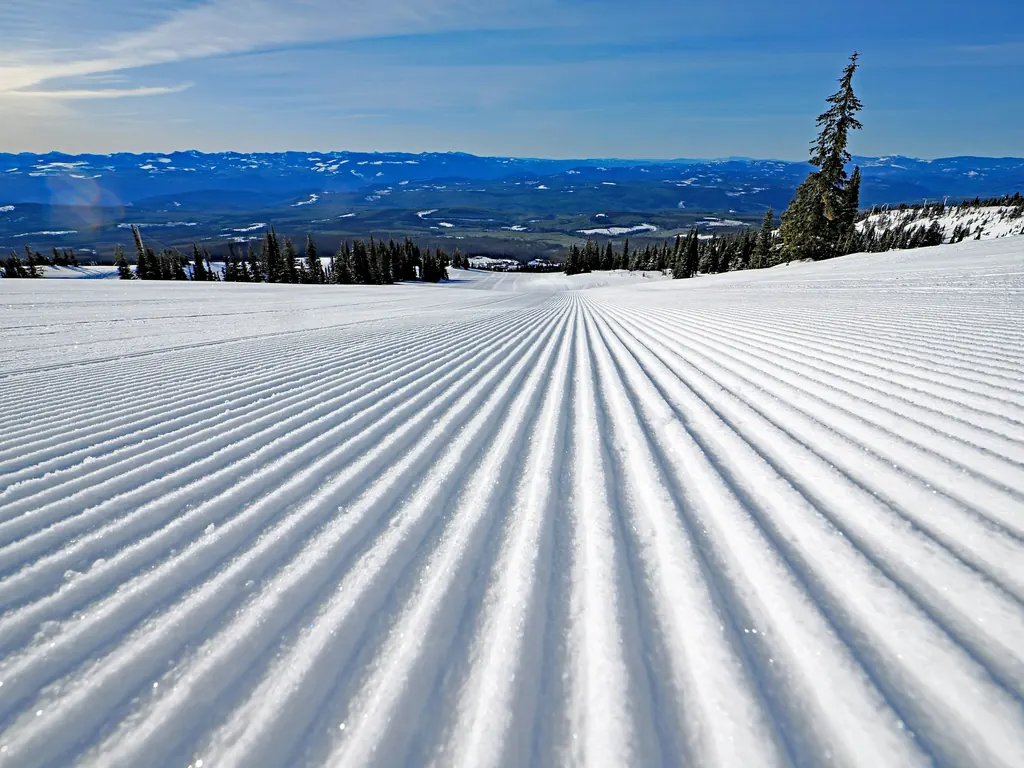 A freshly groomed ski slope in bright sun, looking out over forested valleys